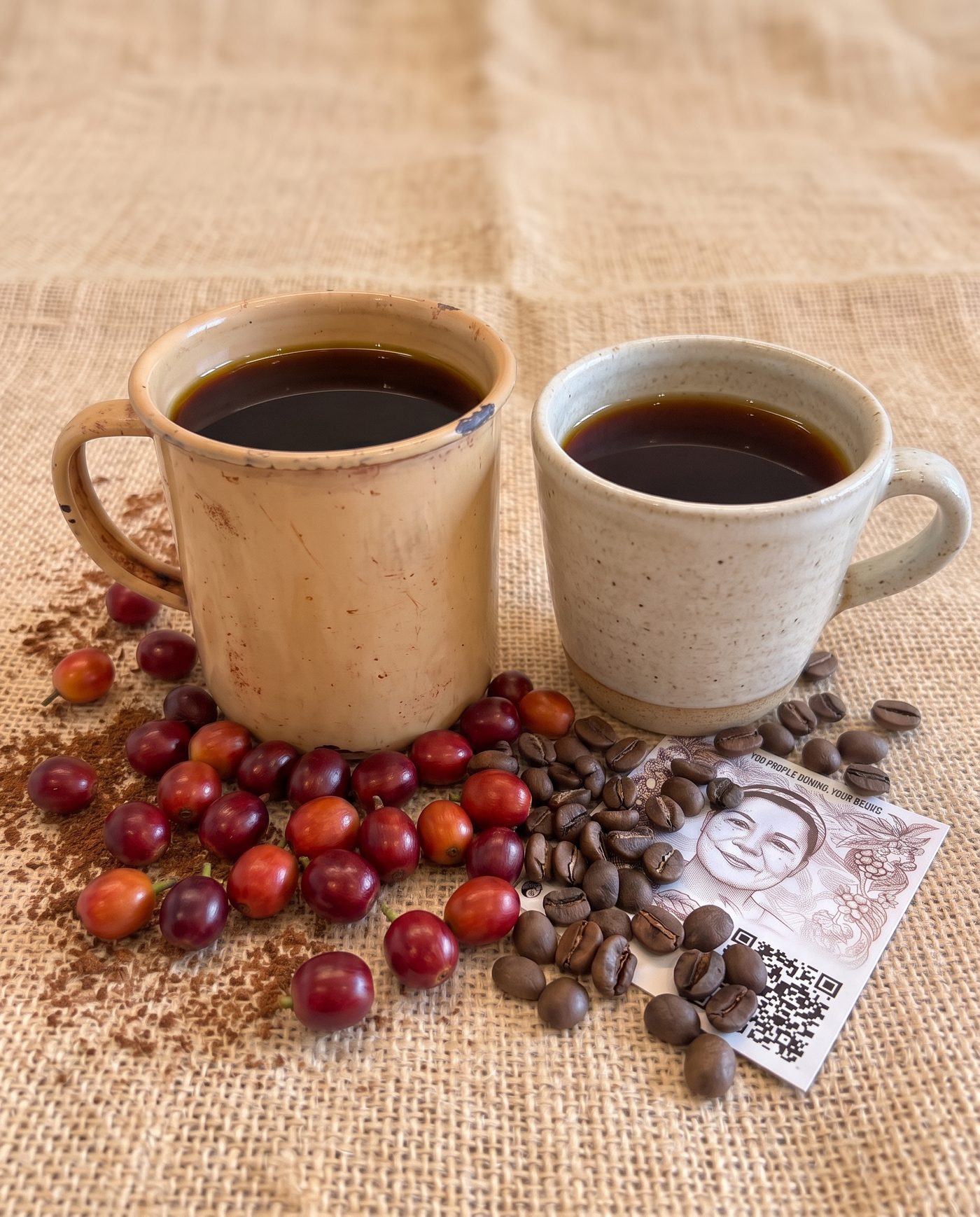 Two ceramic cups of coffee on jute cloth, with coffee cherries, roasted beans and a printed Buna Baraka card beside them.